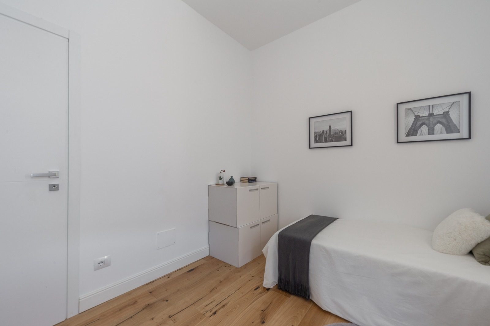 Camera singola con pareti bianche, parquet in rovere, porta bianca e mobili contenitori componibili