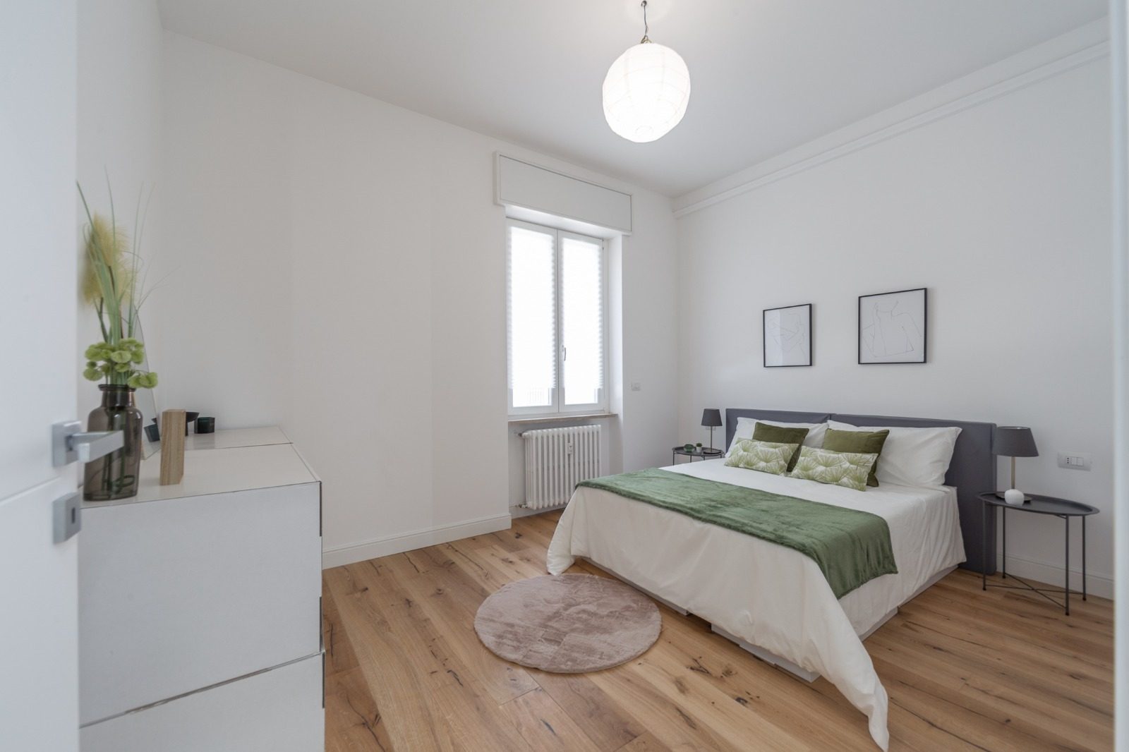 Camera da letto con pareti bianche, pavimento in legno, letto matrimoniale e radiatore sotto la finestra