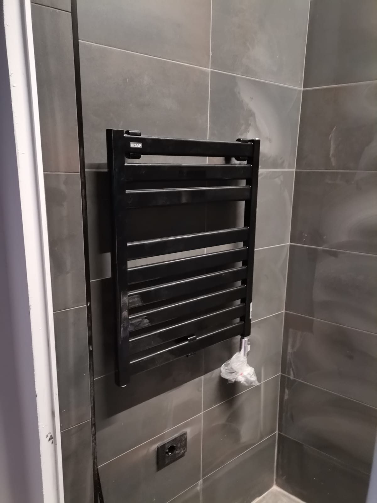 Scaldasalviette nero a parete su rivestimento in gres grigio, cavo elettrico e raccordo ancora scoperti