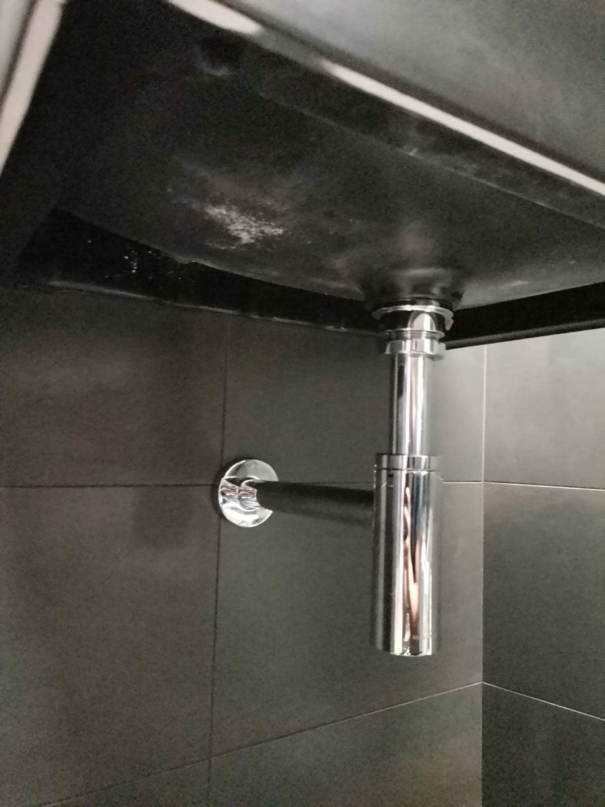 Sifone a bottiglia cromato sotto un lavabo scuro sospeso, parete rivestita in gres grigio