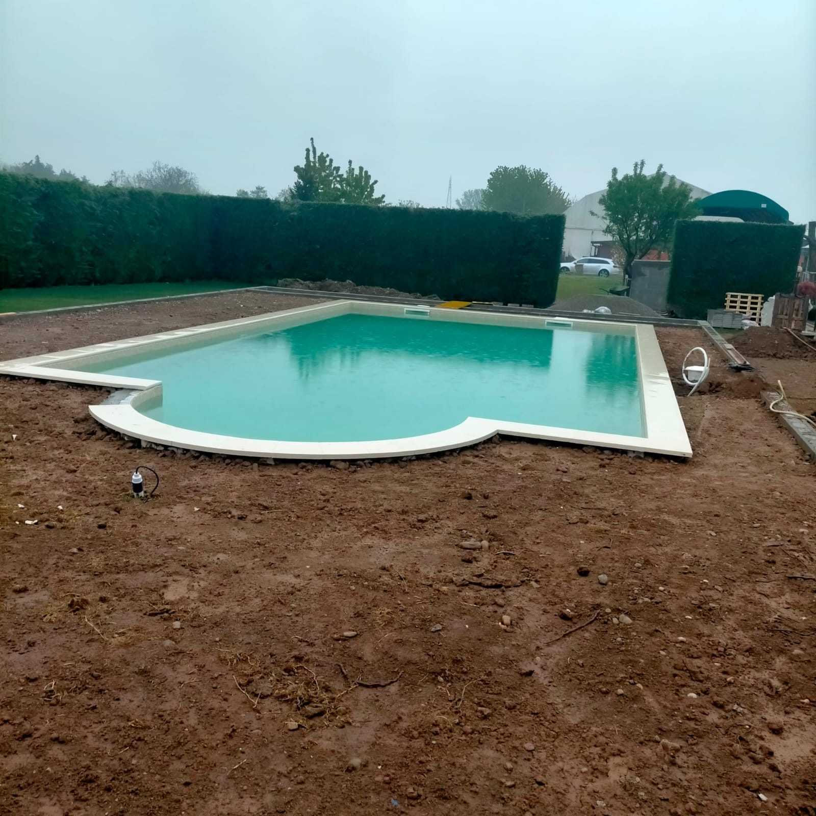 Piscina interrata riempita d'acqua con bordo bianco posato e terreno del giardino ancora da sistemare.