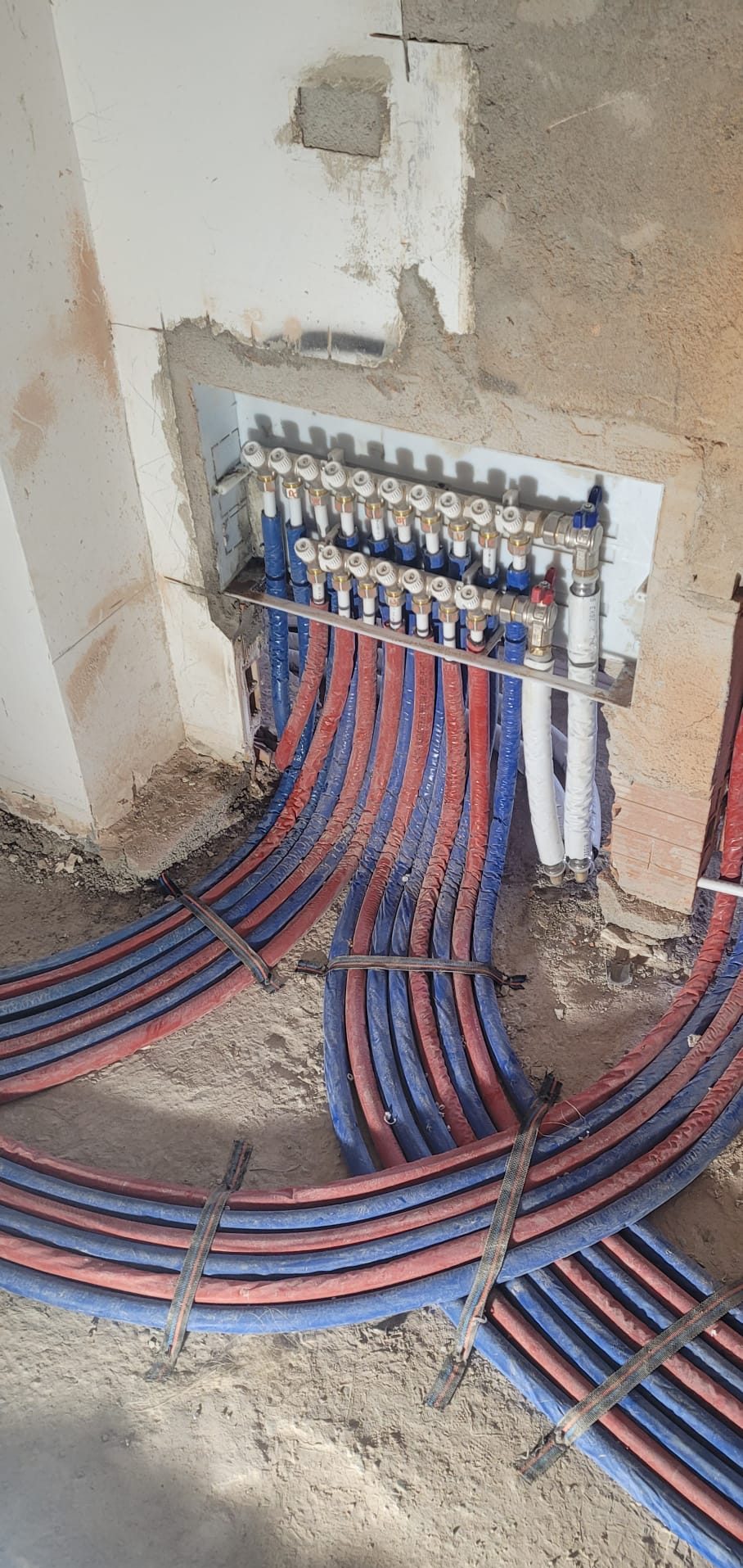 Collettore di distribuzione con valvole incassato nel muro e tubi rossi e blu stesi sul pavimento.