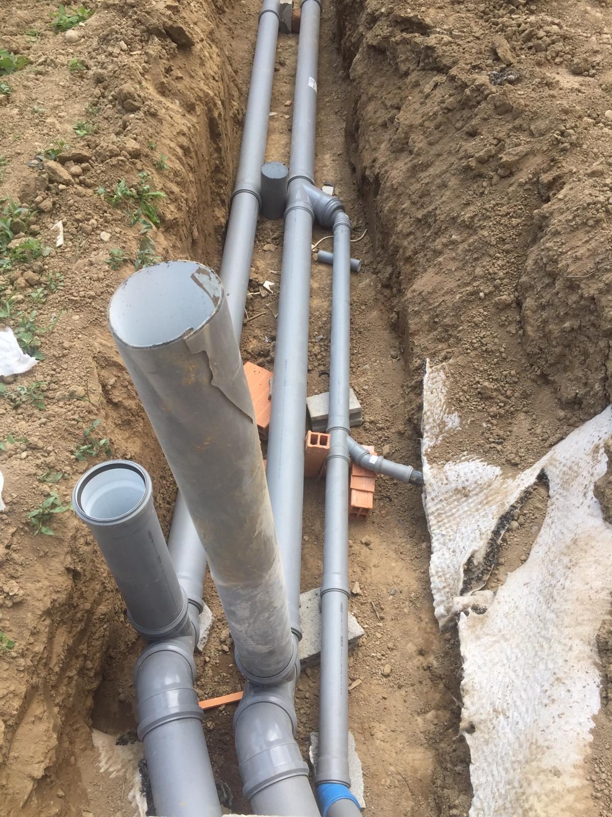 Scavo stretto con tubi di scarico in PVC grigio appoggiati su blocchi, terreno e telo bianco
